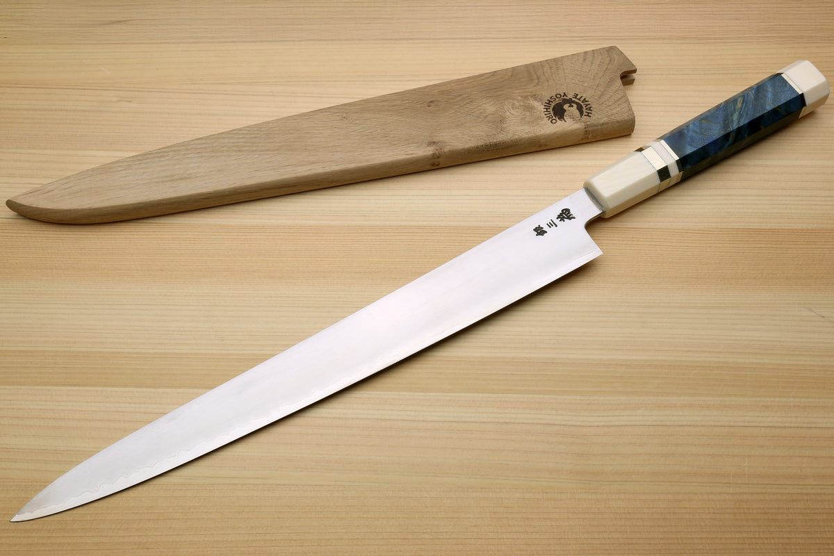 A Japanese-style kitchen knife with a long silver blade and a dark blue marbled handle, displayed diagonally next to its wooden sheath on a light wood grain surface.