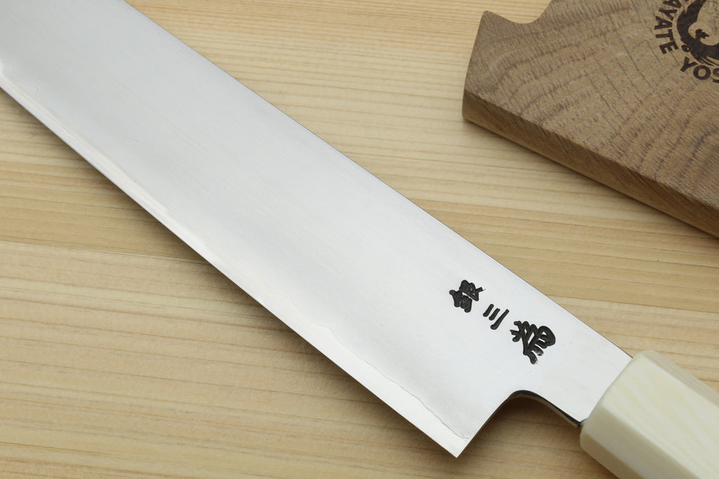A Japanese kitchen knife with a satin-finished silver blade and black kanji characters, resting diagonally on a light wood grain surface next to a wooden block.