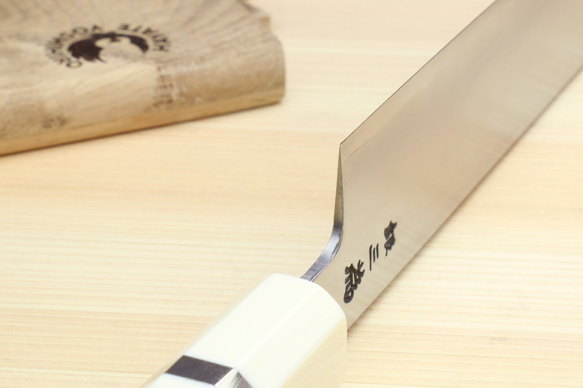 A close-up of a Japanese kitchen knife with a white handle and a silver steel blade, featuring black kanji characters engraved on the blade, resting on a light wood surface.