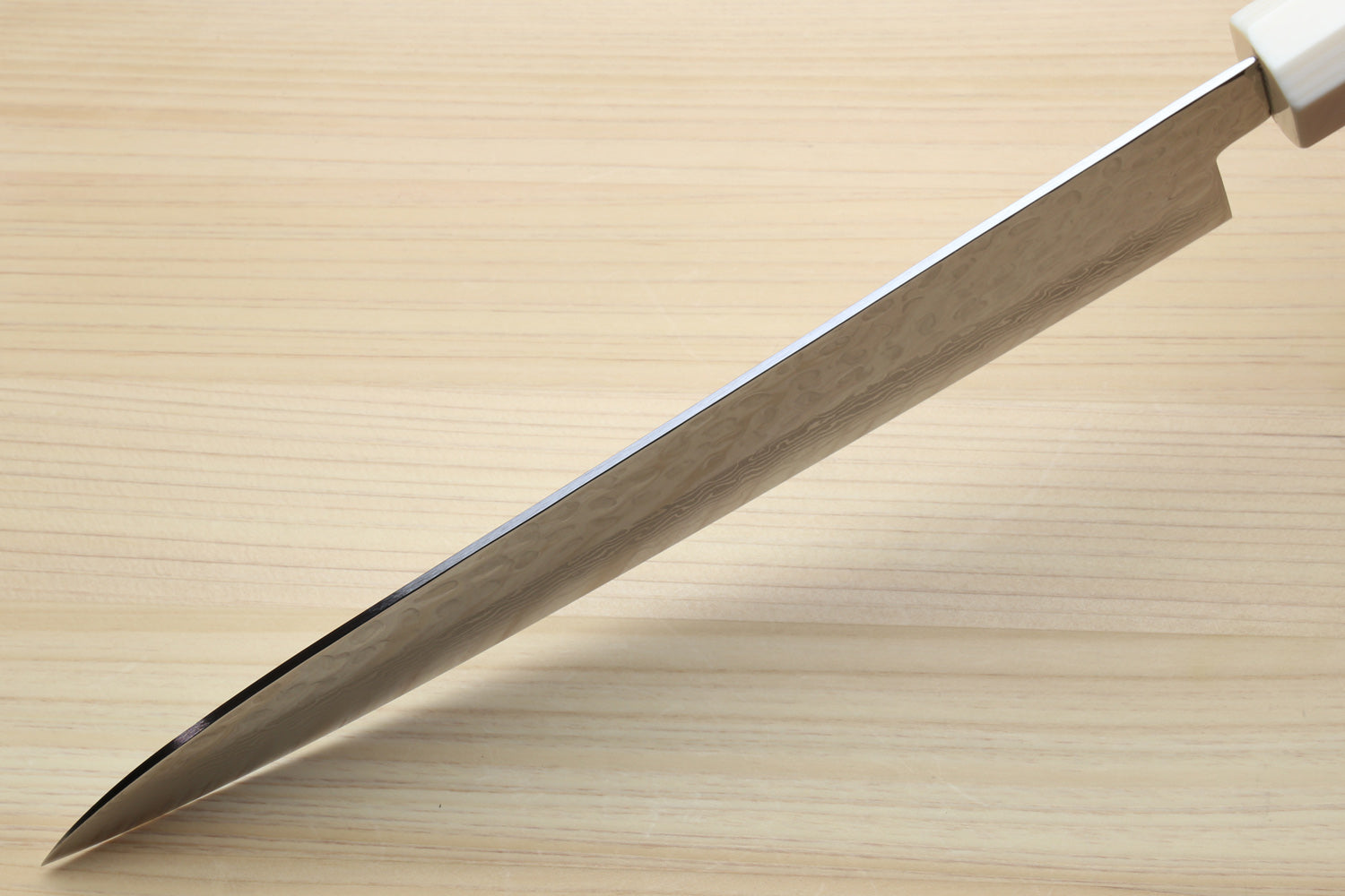 A close-up of a Japanese-style kitchen knife with a satin-finished steel blade and a white handle, resting diagonally on a light wood grain surface.