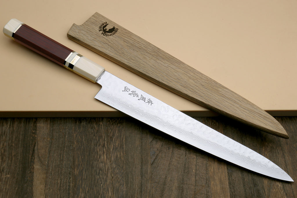 A Japanese-style kitchen knife with a silver patterned blade and a reddish-brown wooden handle, shown diagonally next to its matching wooden sheath on a beige surface with a dark wood tabletop below.