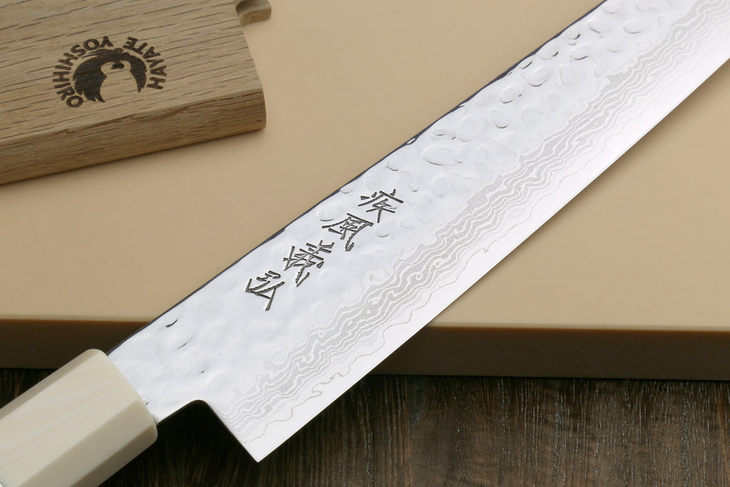 A Japanese kitchen knife with a hammered Damascus steel blade and a cream-colored handle, resting diagonally on a beige background with a wooden box in the upper left corner.