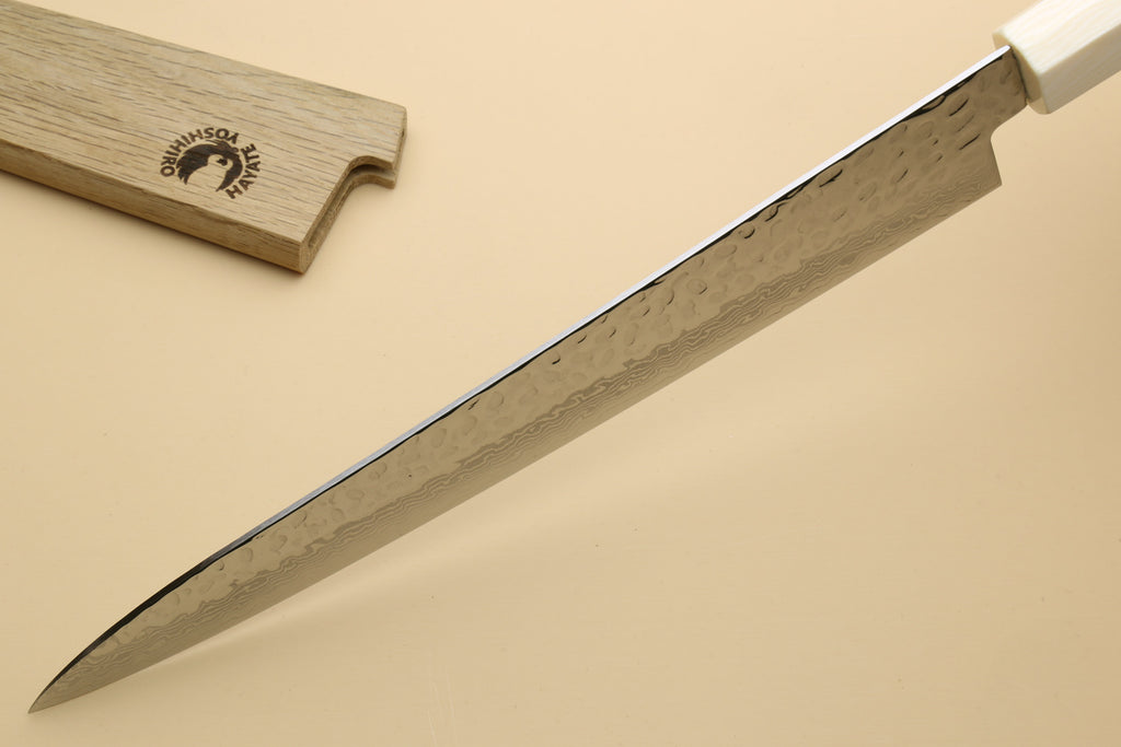 A close-up of a Japanese-style kitchen knife with a hammered Damascus steel blade and a white handle, shown diagonally on a beige background with its wooden box partially visible in the top left.