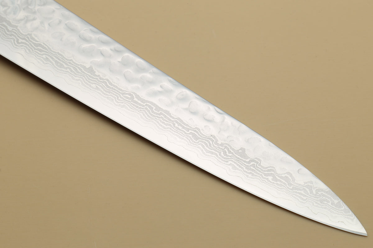 A close-up of a Japanese-style kitchen knife blade with a textured, hammered Damascus steel pattern, set against a solid beige background.
