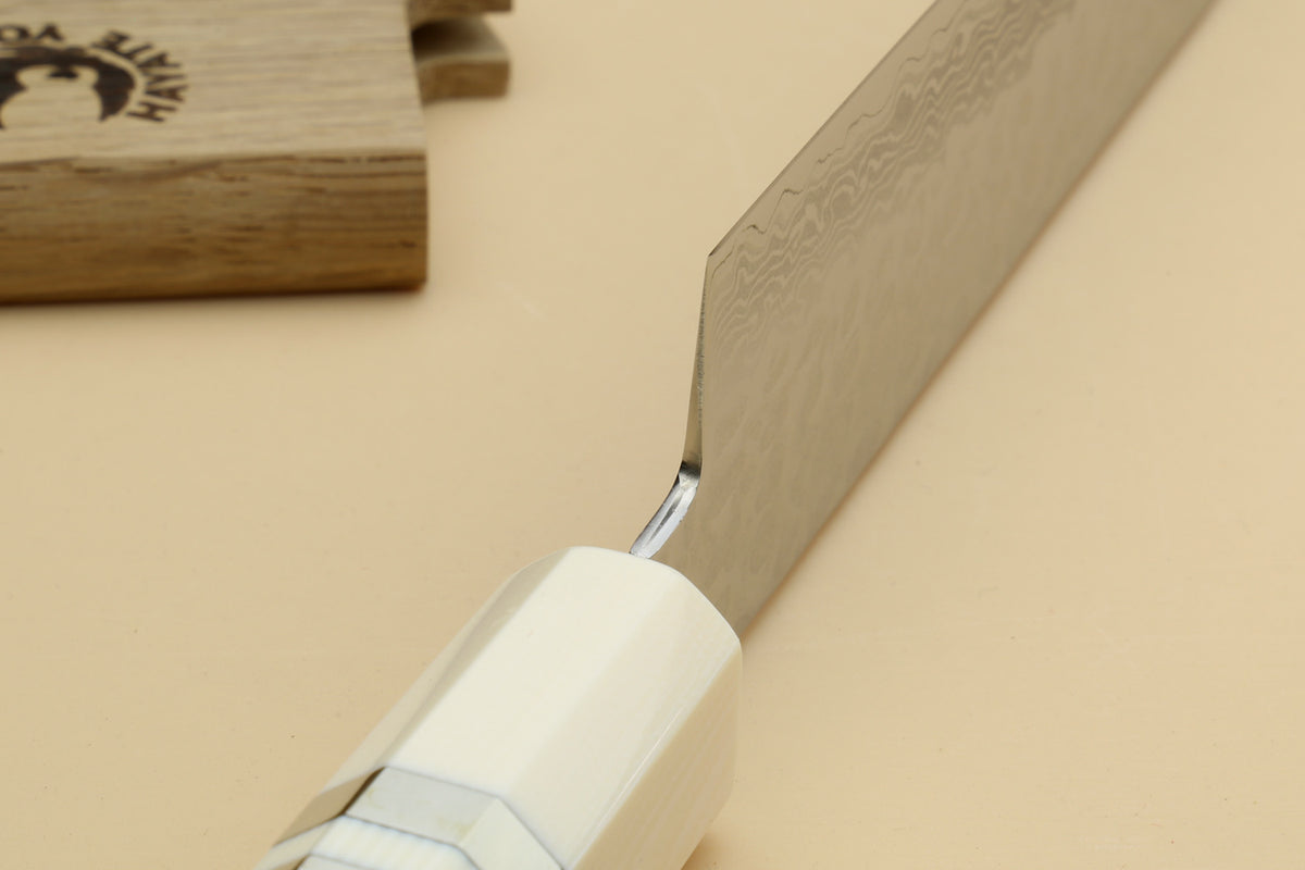 A close-up of a Japanese-style kitchen knife with a Damascus steel blade and a white handle, resting on a beige surface next to its wooden presentation box.