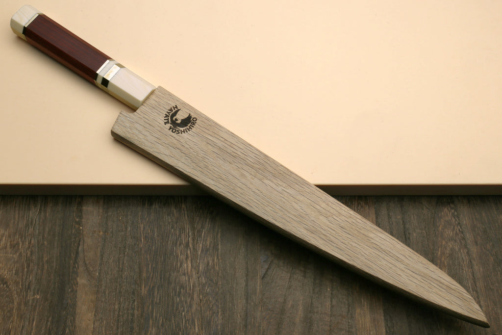 A Japanese-style chef's knife with a gray wood-grain handle and a dark reddish-brown handle end, resting diagonally on a beige surface over a dark wood background. The handle features a black logo with a white bird and the text 'KIKUCHI'.