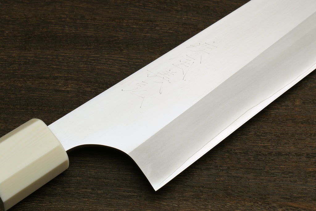 A close-up of a Japanese-style kitchen knife with a satin-finished silver blade and a cream-colored handle, resting on a dark wood surface.