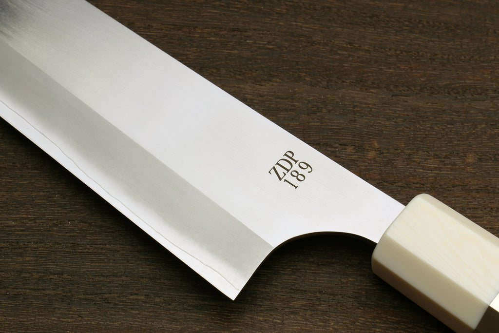 A close-up of a Japanese-style kitchen knife with a satin-finished steel blade and a cream-colored handle, resting on a dark wood surface. The blade is engraved with the brand name 'ZDP 189'.