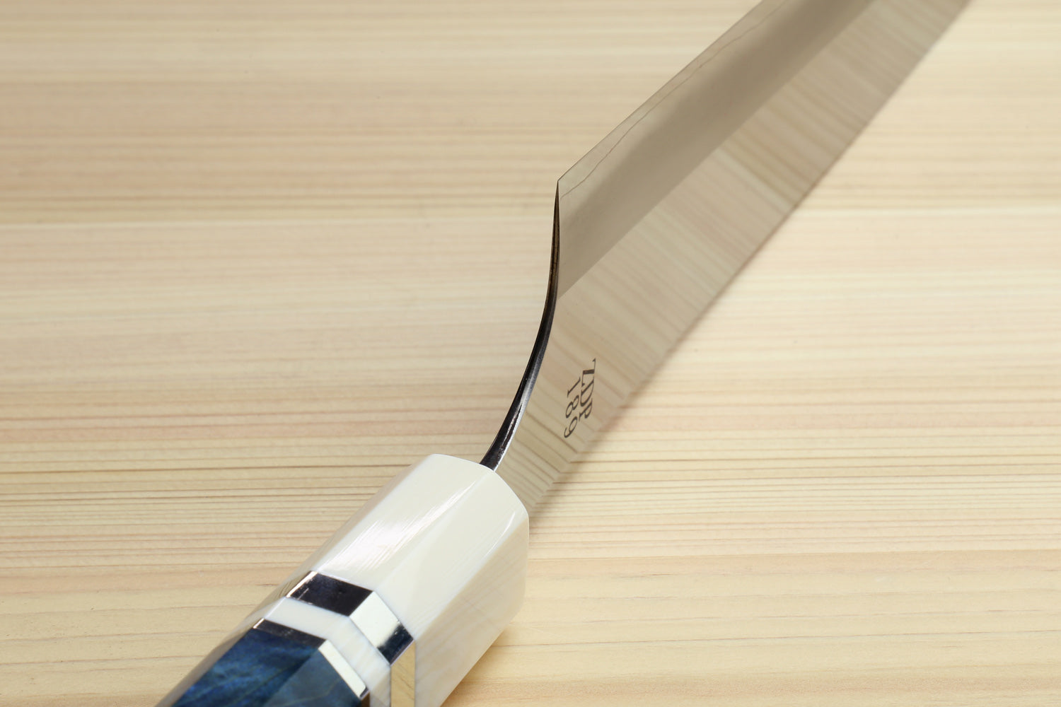 A close-up of a Japanese-style kitchen knife with a satin-finished steel blade and a white handle featuring a dark blue inlay, resting diagonally on a light wood grain surface.