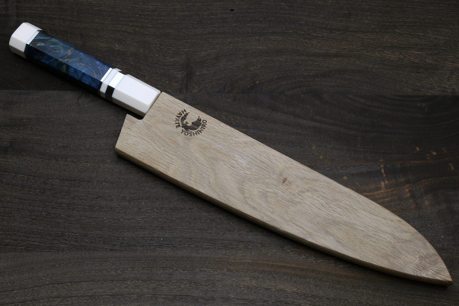 A chef's knife with a natural wood blade and a blue marbled handle, placed diagonally on a dark wood background. The blade features a black logo for 'Black Mountain Knife Co.