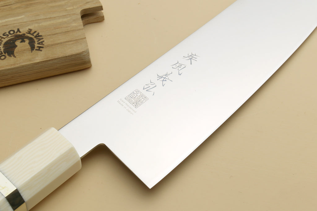 A Japanese-style kitchen knife with a polished silver blade featuring engraved kanji characters and a light-colored wooden handle, displayed diagonally on a beige background with a wooden block in the top-left corner.