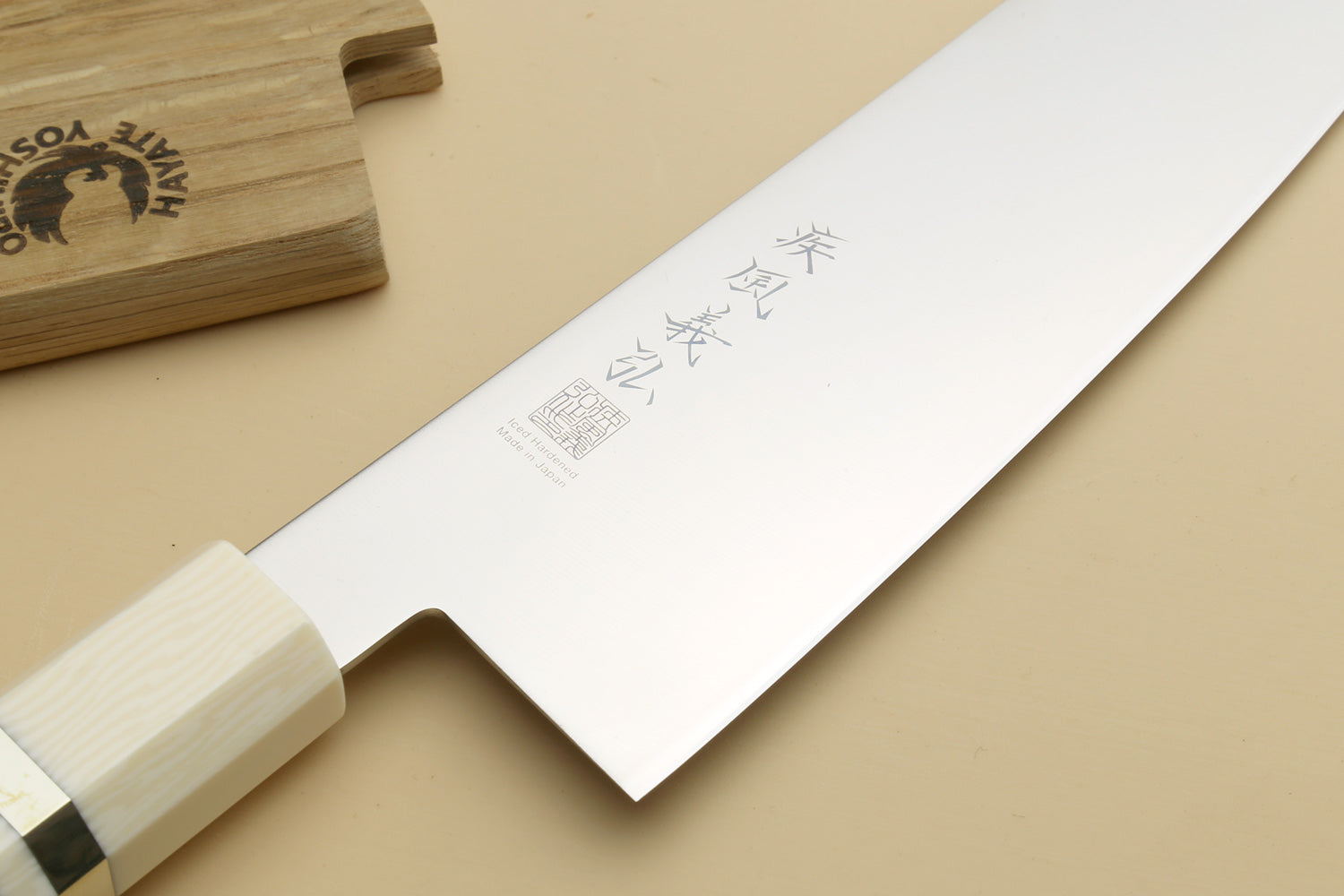A Japanese-style kitchen knife with a polished silver blade featuring engraved kanji characters and a light-colored wooden handle, displayed diagonally on a beige background with a wooden block in the top-left corner.