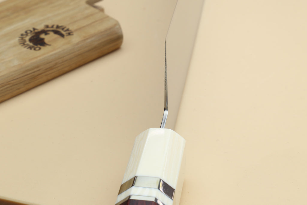 A close-up of a chef's knife with a white octagonal handle and a silver blade, resting on a beige surface next to a wooden cutting board.