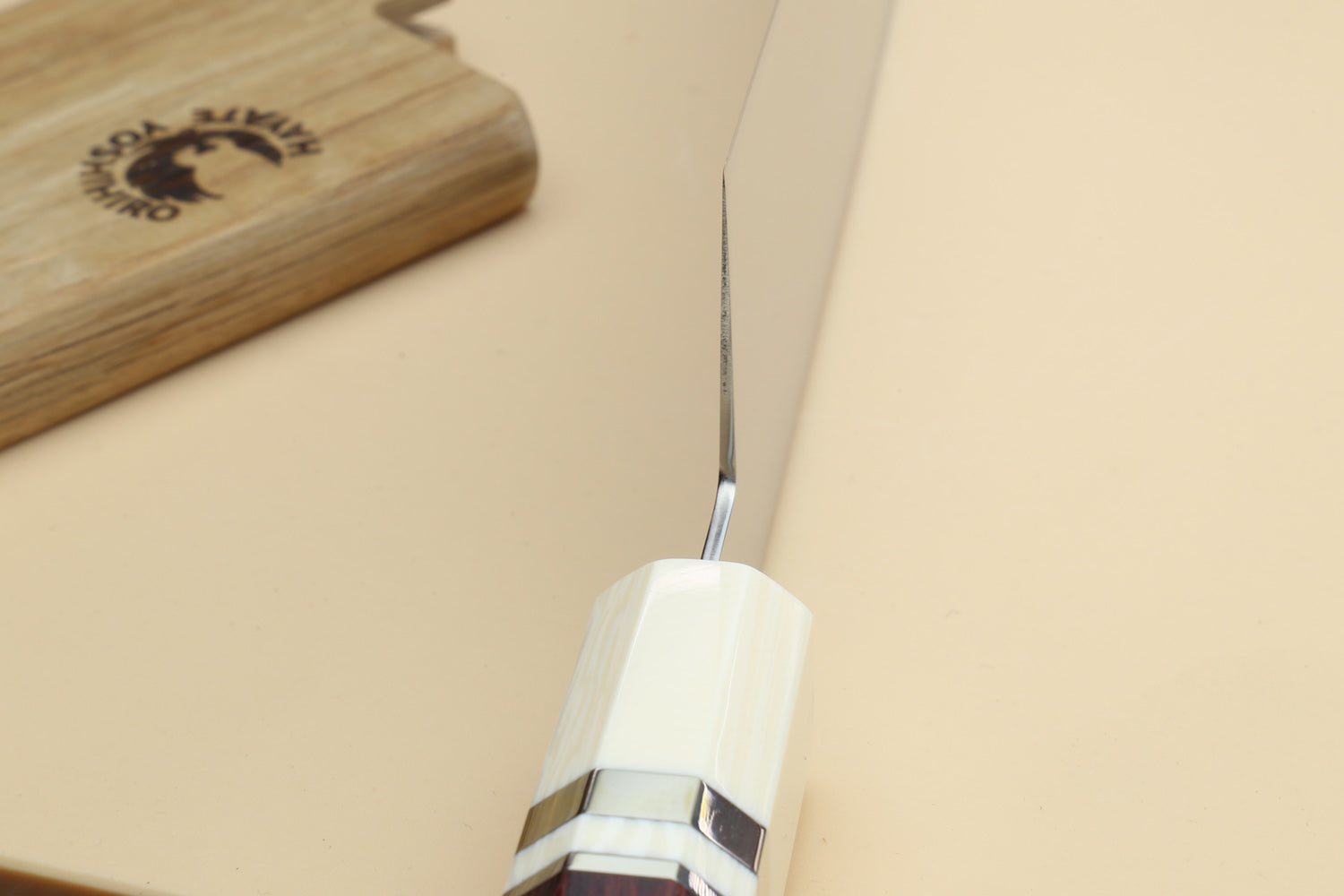 A close-up of a chef's knife with a white octagonal handle and a silver blade, resting on a beige surface next to a wooden cutting board.