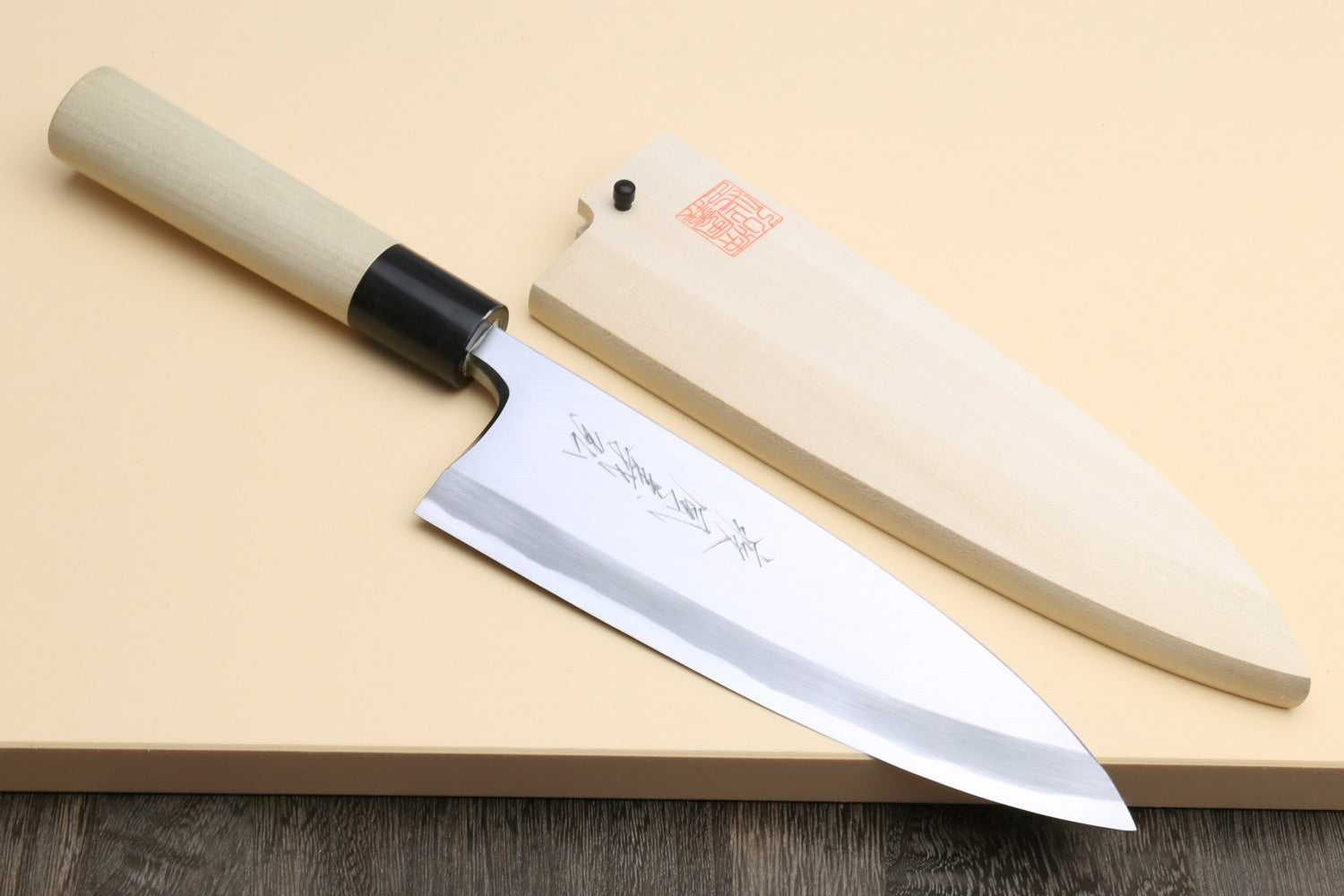 A Japanese-style kitchen knife with a light wood handle and a polished silver blade, shown alongside its matching wooden sheath on a beige surface with a dark wood tabletop visible underneath.
