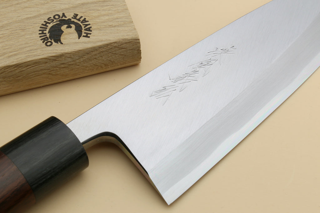 A close-up of a Japanese-style kitchen knife with a polished silver blade, etched kanji characters, a dark handle, and a wooden box branded HAITE YOSHIO in the background.
