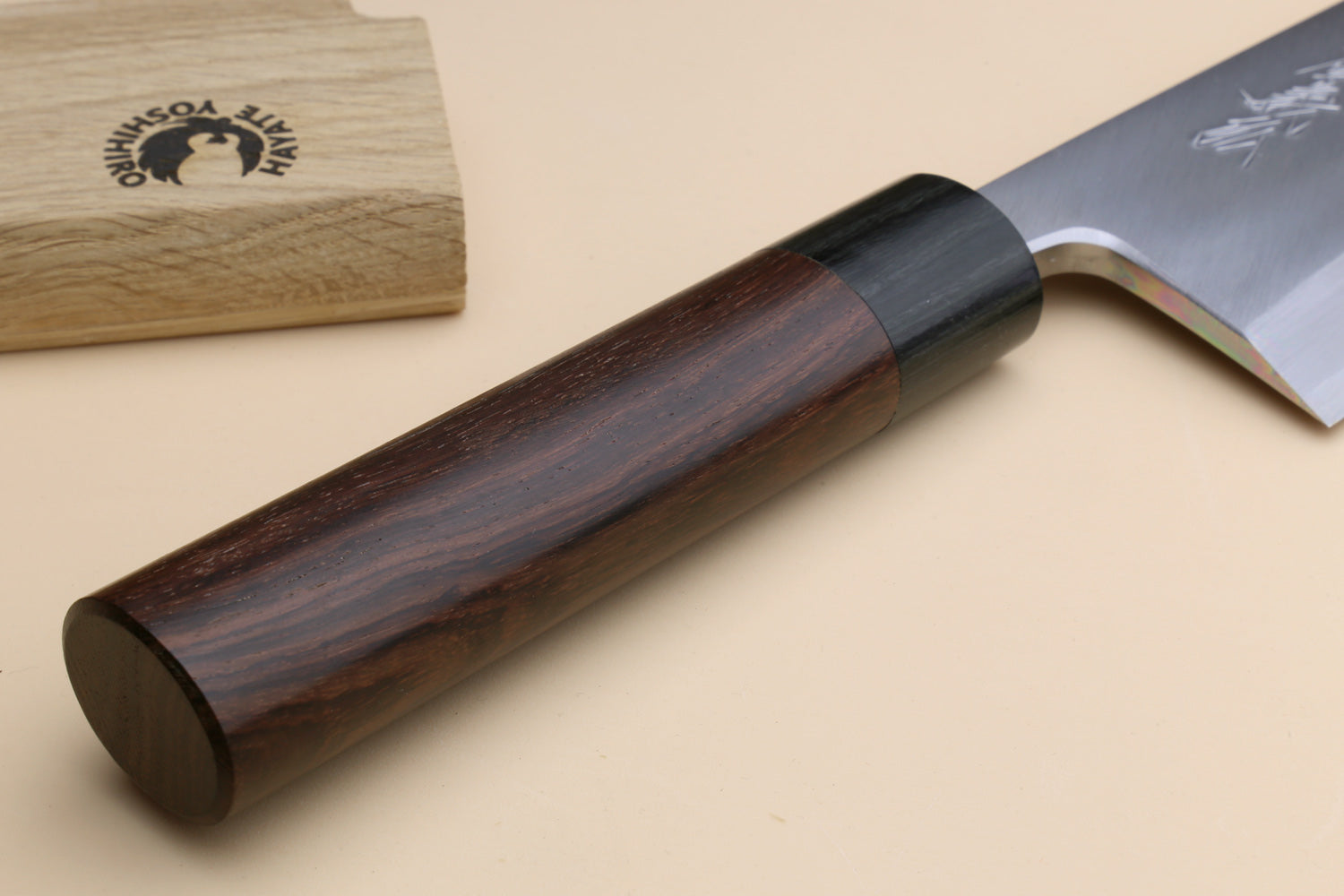 A close-up of a Japanese-style kitchen knife with a dark brown wood handle and a silver steel blade, resting on a beige surface next to a branded wooden box.