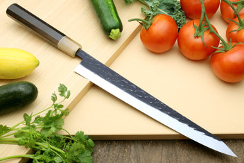 Stainless chef knife with speckled blade and dark handle, with tomatoes, zucchini, and herbs