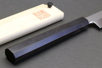 Sushi chef knife with mirror blade and dark ebony handle, resting on light saya.