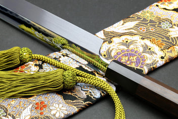 Japanese knife with silver blade and black handle beside green tassel cord and patterned cloth.