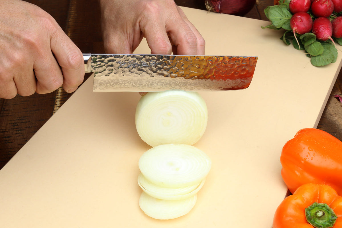 Petty knife with hammered Damascus blade cutting onion on a cutting board.